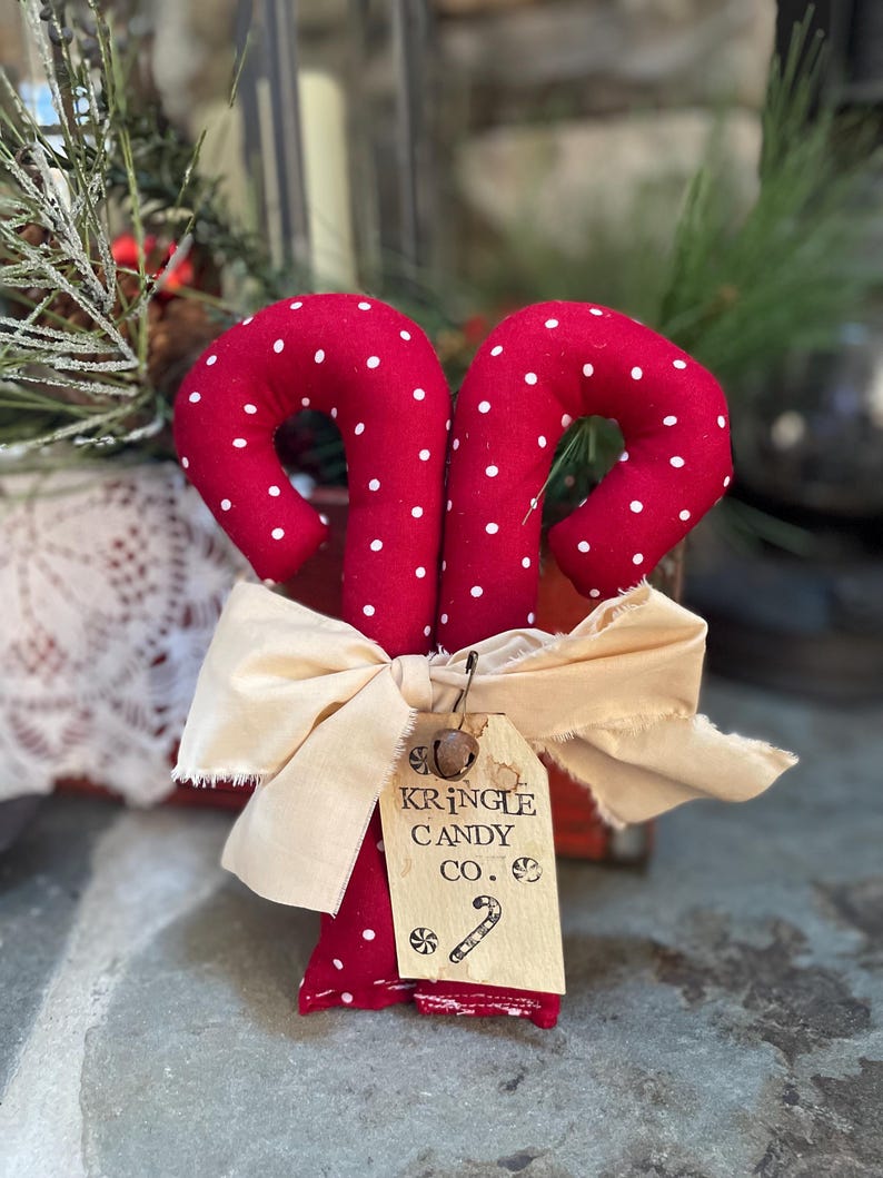 May include: Two red fabric candy canes with white polka dots are tied together with a cream-colored bow. A small tag reads "KRINGLE CANDY CO." with a candy cane illustration. Festive holiday decor.
