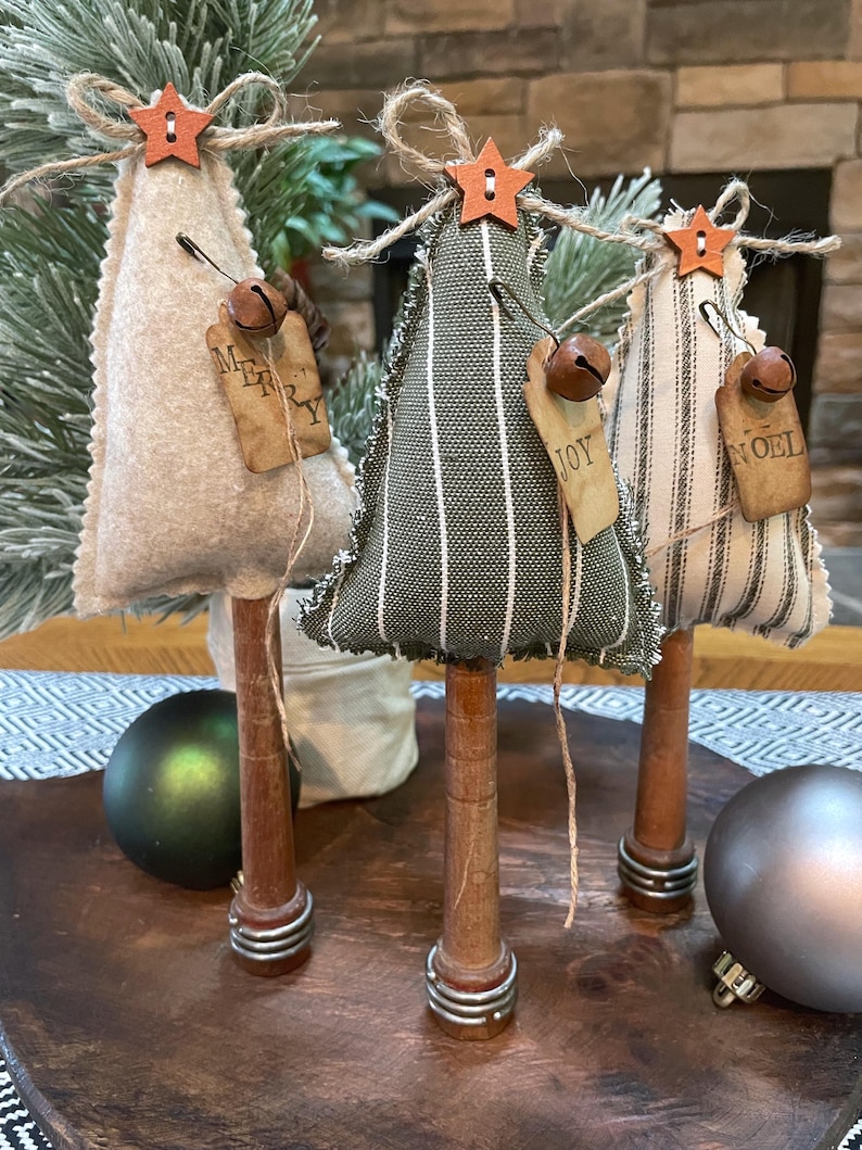 May include: Three miniature Christmas trees made of fabric with a rustic wood base. The trees are decorated with burlap ribbon, a star, and a bell. The trees are labeled "Merry", "Joy", and "Noel".