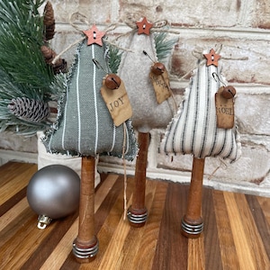 May include: Three handmade fabric Christmas trees with striped fabric, wooden bases, and metal rings. The trees are decorated with bells, stars, and tags that say "Joy", "Merry", and "Noel".
