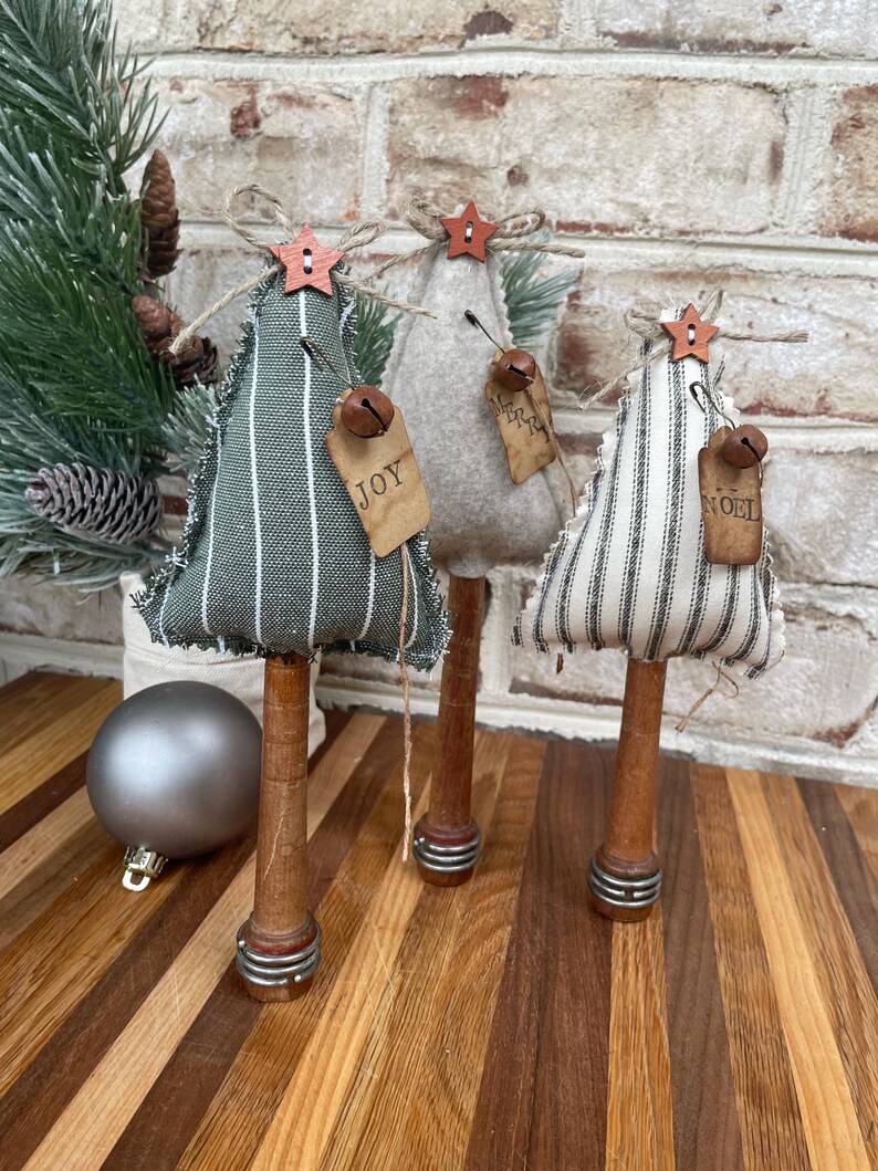 May include: Three handmade fabric Christmas trees with striped fabric, wooden bases, and metal rings. The trees are decorated with bells, stars, and tags that say "Joy", "Merry", and "Noel".