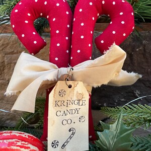May include: Two red fabric candy canes with white polka dots, tied together with a cream-colored bow. A tag reads "Kringle Candy Co." with a candy cane illustration. Festive holiday decor.