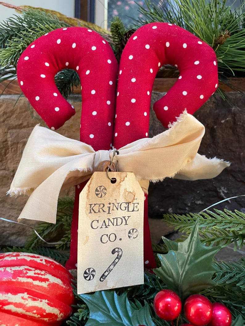 May include: Two red fabric candy canes with white polka dots, tied together with a cream-colored bow. A tag reads "Kringle Candy Co." with a candy cane illustration. Festive holiday decor.