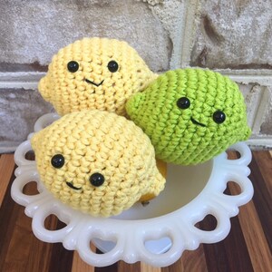 May include: Three crocheted fruit plushies: two yellow lemons and one green lime. Each plush has a smiling face with black eyes and a stitched mouth.