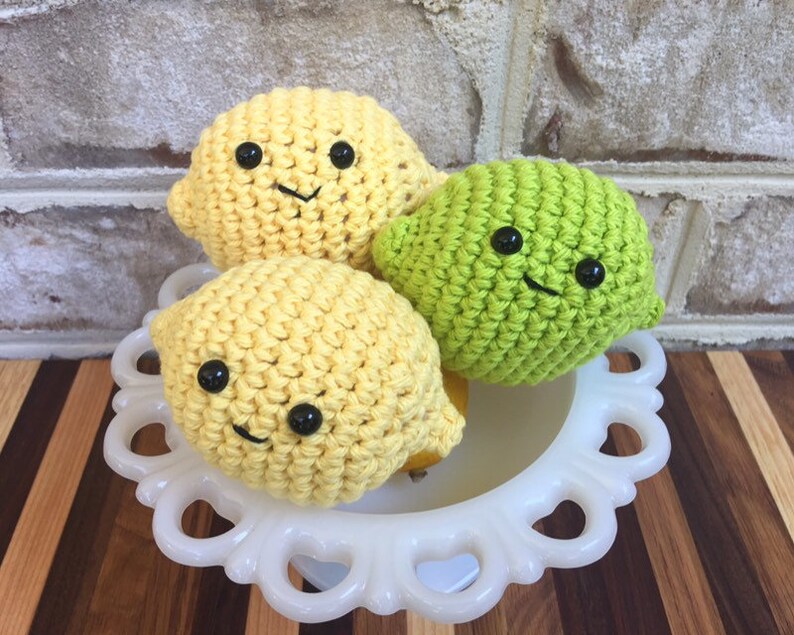 May include: Three crocheted fruit plushies: two yellow lemons and one green lime. Each plush has a smiling face with black eyes and a stitched mouth.