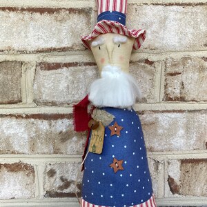 K&ouml;nnte beinhalten: Eine handgemachte patriotische Puppe, die einen rot-wei&szlig;-gestreiften Hut, ein blau-wei&szlig; gepunktetes Kleid und einen rot-wei&szlig;-gestreiften Rock tr&auml;gt. Die Puppe hat einen wei&szlig;en Bart und ein Schild mit der Aufschrift "1776".