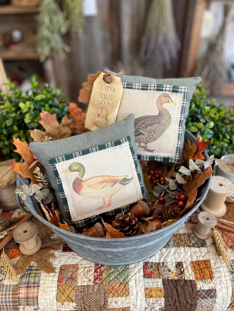 May include: Two square fabric pillows in a galvanized metal basin. One pillow features a mallard duck, the other a brown duck. Both have green plaid borders. A tan tag attached to the pillows reads, "INTO THE WOODS I GO". The basin contains autumn leaves.