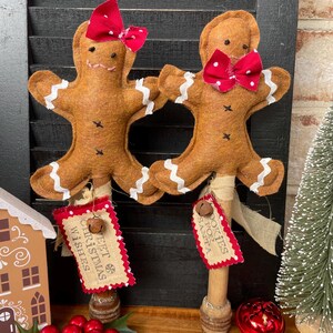 May include: Two gingerbread man ornaments with red bow ties and white icing details. Each ornament has a tag that says "Sweet Christmas Wishes" and "Cookies for Santa". The ornaments are on wooden sticks.