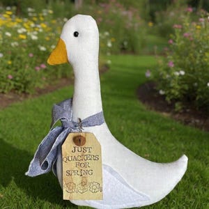 May include: A white fabric goose with a yellow beak and a gray ribbon tied around its neck. A wooden tag with the words "Just Quackers for Spring" is attached. The goose is set against a backdrop of green grass and a flower garden.