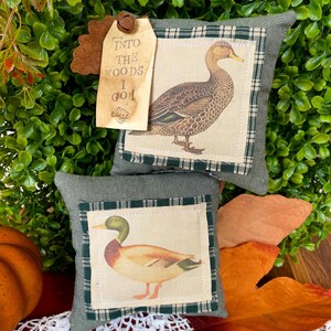 May include: Two square olive green fabric pillows featuring centered cream-colored patches with illustrations of ducks. A tan paper tag attached with a small metal bell reads, "INTO THE WOODS I GO!" with a small oak leaf icon at the bottom.