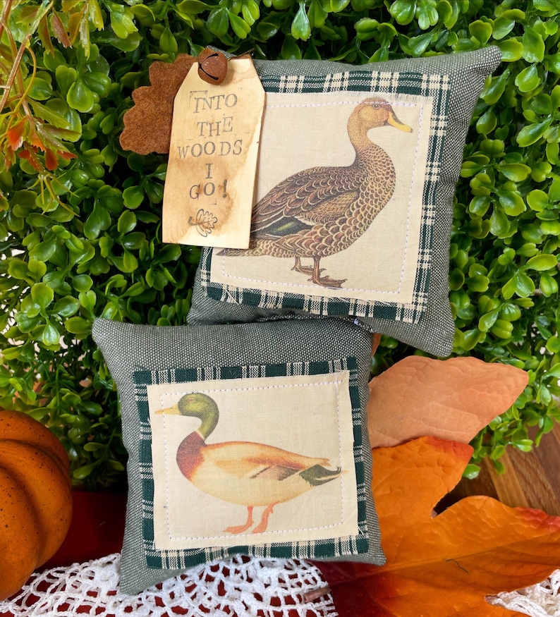 May include: Two square olive green fabric pillows featuring centered cream-colored patches with illustrations of ducks. A tan paper tag attached with a small metal bell reads, "INTO THE WOODS I GO!" with a small oak leaf icon at the bottom.