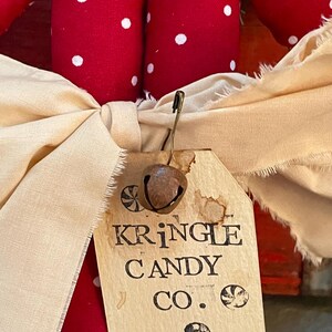 May include: Close-up of a Christmas decoration featuring red fabric with white polka dots. A cream-colored bow is tied around the decoration, with a tag that reads "KRINGLE CANDY CO." and a small bell attached.