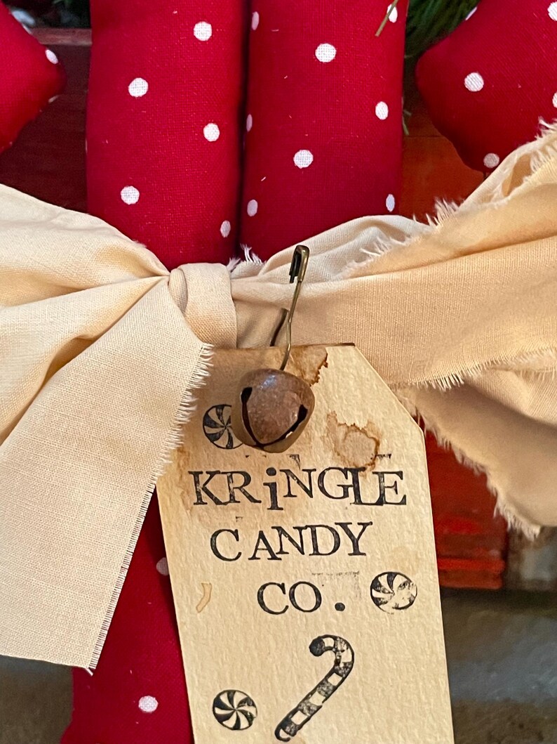 May include: Close-up of a Christmas decoration featuring red fabric with white polka dots. A cream-colored bow is tied around the decoration, with a tag that reads "KRINGLE CANDY CO." and a small bell attached.
