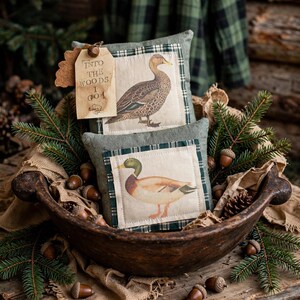 May include: Two small square pillows featuring duck illustrations on cream fabric with green plaid borders. A tan paper tag attached to the top pillow displays the text "INTO THE WOODS I GO" with a small acorn graphic printed in black ink.