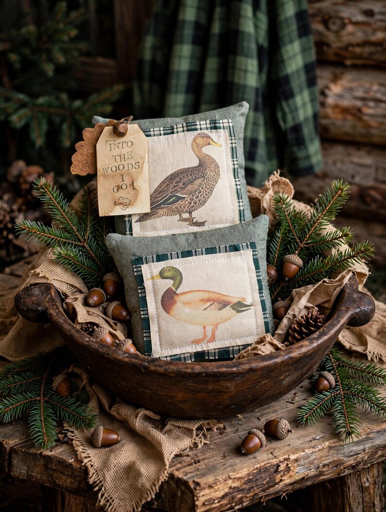 May include: Two small square pillows featuring duck illustrations on cream fabric with green plaid borders. A tan paper tag attached to the top pillow displays the text "INTO THE WOODS I GO" with a small acorn graphic printed in black ink.