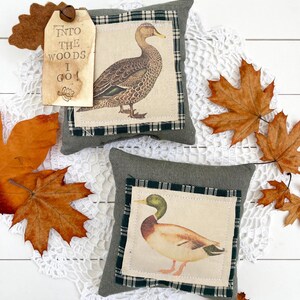 May include: Two square olive green fabric pillows featuring centered patches with duck illustrations. One pillow has a tan tag attached with a small metal bell, printed with the text "INTO THE WOODS I GO!" and a small leaf graphic.