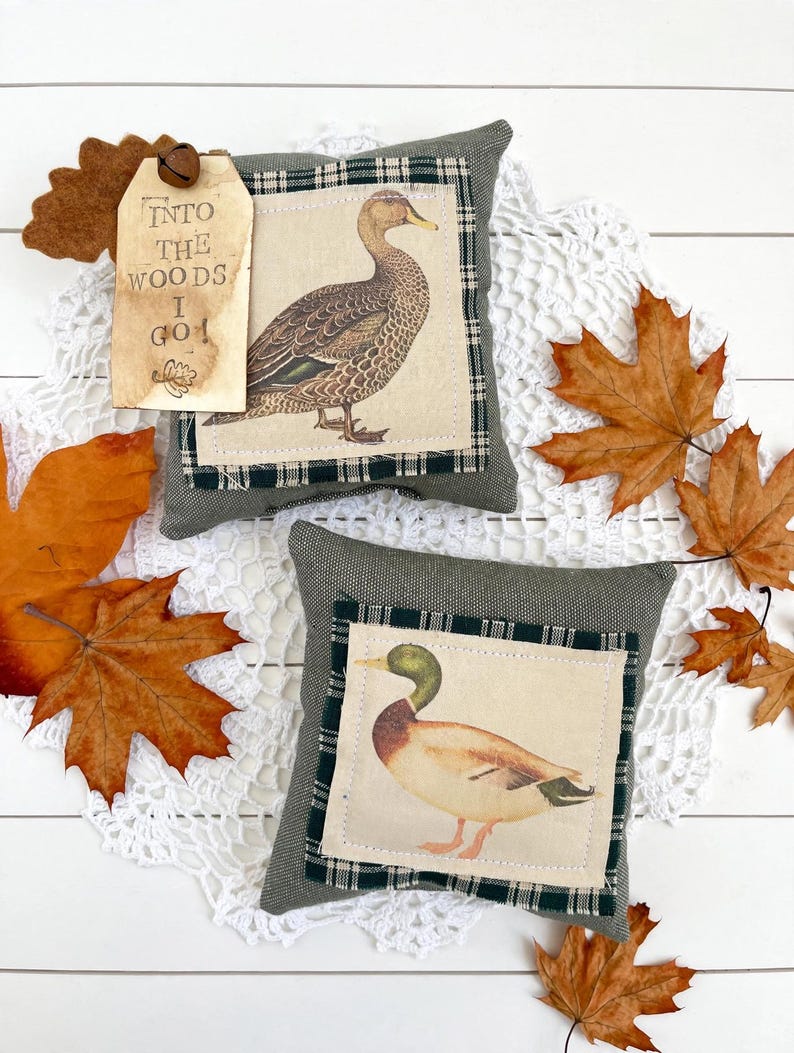 May include: Two square olive green fabric pillows featuring centered patches with duck illustrations. One pillow has a tan tag attached with a small metal bell, printed with the text "INTO THE WOODS I GO!" and a small leaf graphic.