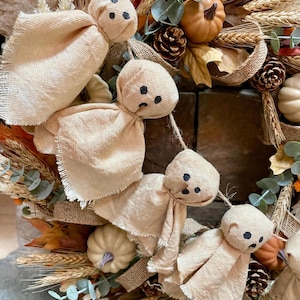 May include: A decorative garland featuring four small ghost figures made of light beige fabric with hand-drawn black facial features, suspended on a string and nestled within a wreath of dried wheat, pinecones, eucalyptus, and small pumpkins.