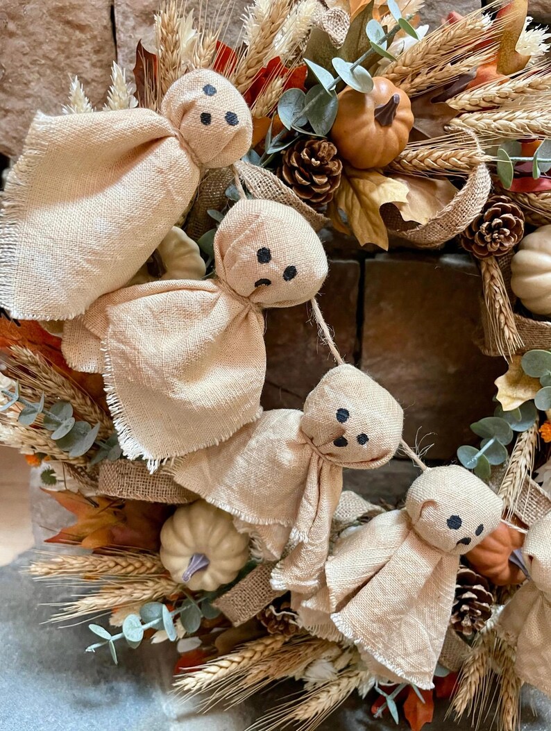 May include: A decorative garland featuring four small ghost figures made of light beige fabric with hand-drawn black facial features, suspended on a string and nestled within a wreath of dried wheat, pinecones, eucalyptus, and small pumpkins.