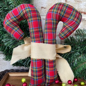 May include: Two fabric candy canes featuring a red, yellow, and blue plaid pattern, tied together with a wide, frayed-edge beige ribbon. The candy canes are stuffed and have a curved top, positioned upright as a decorative holiday ornament.
