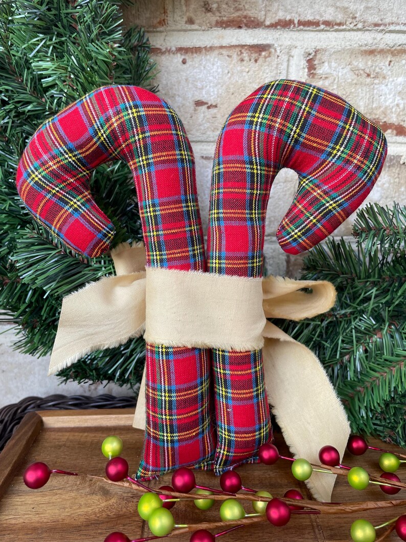 May include: Two fabric candy canes featuring a red, yellow, and blue plaid pattern, tied together with a wide, frayed-edge beige ribbon. The candy canes are stuffed and have a curved top, positioned upright as a decorative holiday ornament.