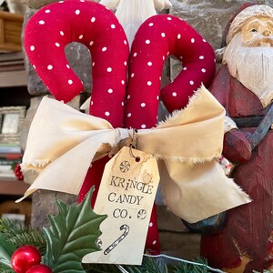 May include: Two red fabric candy canes with white polka dots are tied together with a cream-colored bow. A tag reads "Kringle Candy Co." with a candy cane illustration. Christmas decor.