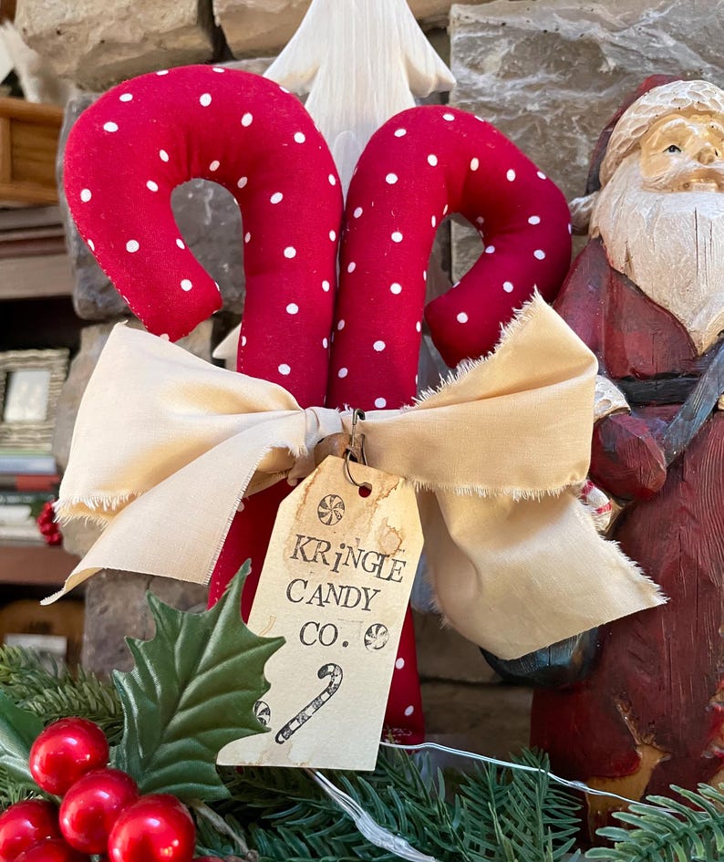May include: Two red fabric candy canes with white polka dots are tied together with a cream-colored bow. A tag reads "Kringle Candy Co." with a candy cane illustration. Christmas decor.