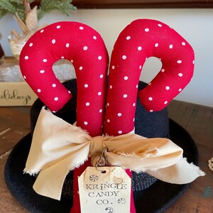 May include: Red fabric candy canes with white polka dots, resting on a black hat. A cream-colored bow and a tag that reads "Kringle Candy Co." are attached. The candy canes are a festive holiday decoration.