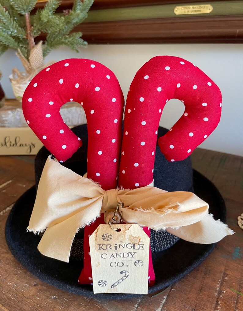 May include: Red fabric candy canes with white polka dots, resting on a black hat. A cream-colored bow and a tag that reads "Kringle Candy Co." are attached. The candy canes are a festive holiday decoration.