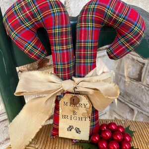 May include: Two fabric candy canes featuring a red, green, yellow, and blue plaid pattern. A beige frayed ribbon is tied around the center, holding a rectangular paper tag with the text MERRY & BRIGHT! and a small metal bell attached.