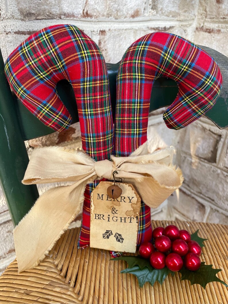May include: Two fabric candy canes featuring a red, green, yellow, and blue plaid pattern. A beige frayed ribbon is tied around the center, holding a rectangular paper tag with the text MERRY & BRIGHT! and a small metal bell attached.