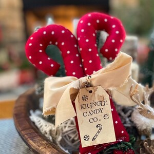 May include: Two red candy canes with white polka dots, tied with a beige ribbon and a tag that reads "Kringle Candy Co." The candy canes are in a wooden bowl with other Christmas decorations.
