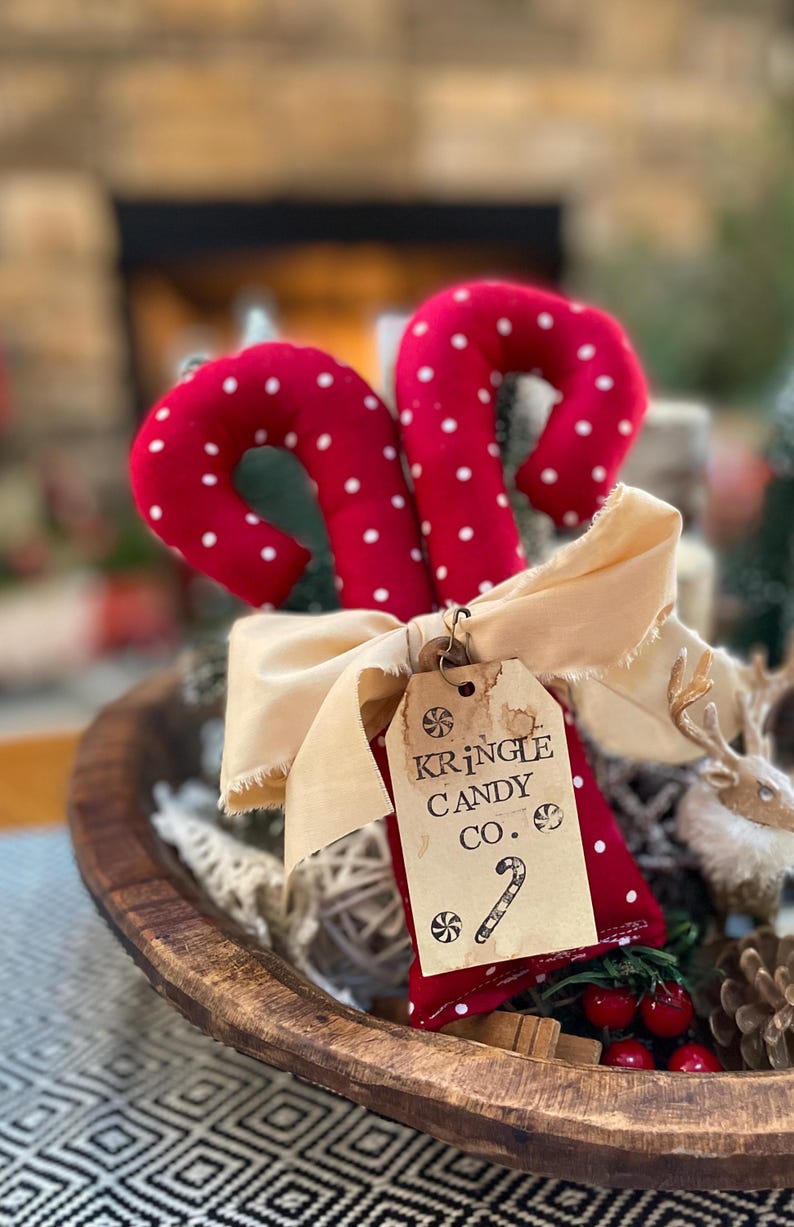 May include: Two red candy canes with white polka dots, tied with a beige ribbon and a tag that reads "Kringle Candy Co." The candy canes are in a wooden bowl with other Christmas decorations.