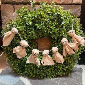 May include: A circular green boxwood wreath featuring a hanging garland of six small, beige fabric ghost figures. Each ghost is crafted from light-colored cloth with frayed edges and features simple black dots for eyes and a mouth, strung on twine.