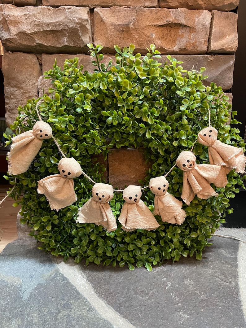 May include: A circular green boxwood wreath featuring a hanging garland of six small, beige fabric ghost figures. Each ghost is crafted from light-colored cloth with frayed edges and features simple black dots for eyes and a mouth, strung on twine.