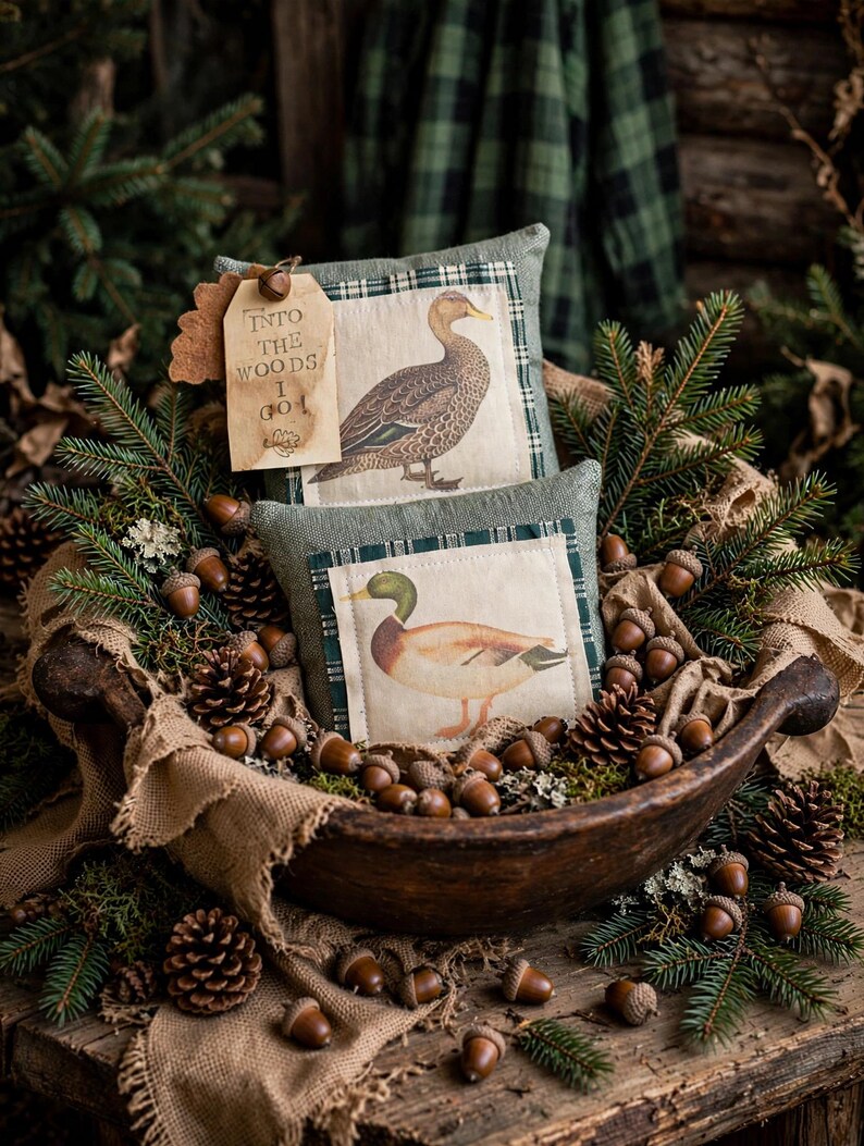 May include: Two square fabric pillows featuring illustrations of ducks, nestled in a dark wooden bowl with pine branches, pinecones, and acorns. A tan paper tag attached to the top pillow displays the text "INTO THE WOODS I GO".