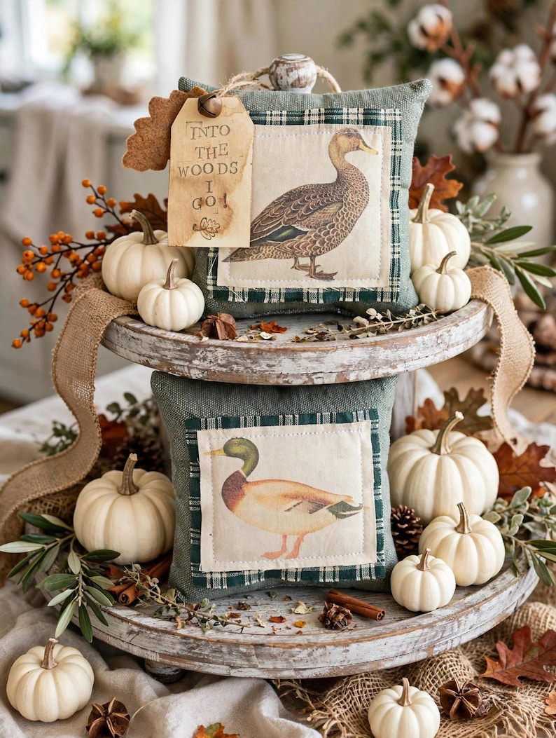 May include: Two square sage green fabric pillows featuring duck illustrations on cream-colored patches. The top pillow has a tag with the text INTO THE WOODS I GO. The pillows are displayed on a two-tiered wooden tray surrounded by white pumpkins.