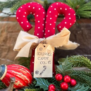 May include: A pair of red fabric candy canes with white polka dots, tied with a cream-colored ribbon. A tag reads "Kringle Candy Co." with a candy cane illustration. Festive holiday decor.