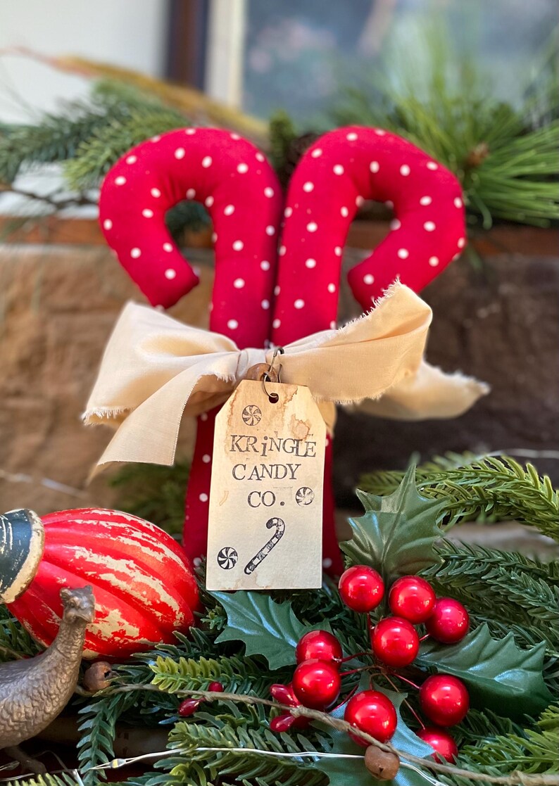 May include: A pair of red fabric candy canes with white polka dots, tied with a cream-colored ribbon. A tag reads "Kringle Candy Co." with a candy cane illustration. Festive holiday decor.