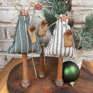 May include: Three miniature Christmas trees made of fabric with striped patterns. The trees are decorated with wooden stars, bells, and fabric tags that say "Joy", "Merry", and "Noel". The trees are mounted on wooden dowels with metal accents.