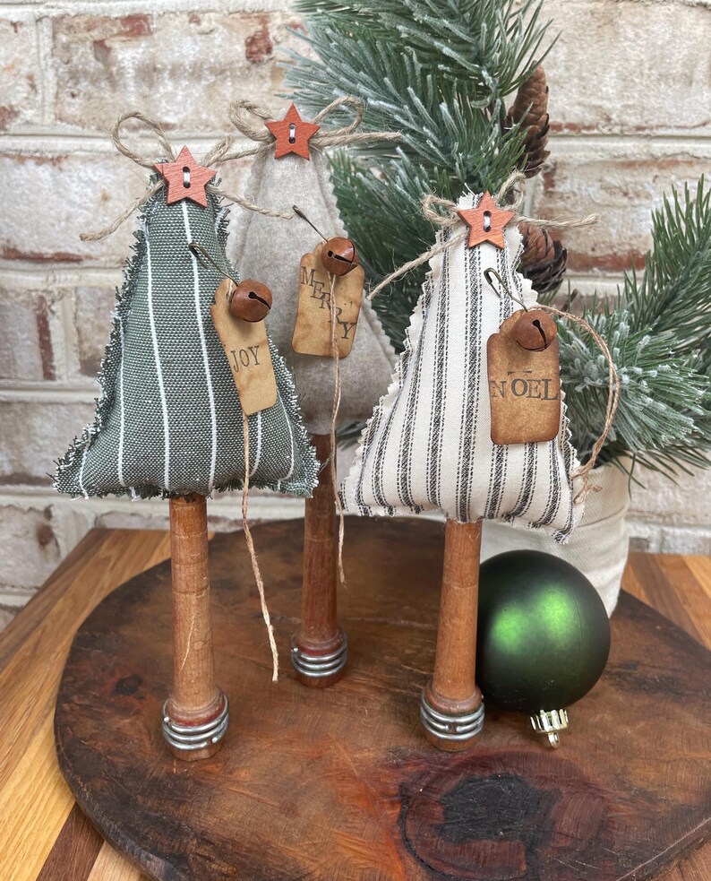May include: Three miniature Christmas trees made of fabric with striped patterns. The trees are decorated with wooden stars, bells, and fabric tags that say "Joy", "Merry", and "Noel". The trees are mounted on wooden dowels with metal accents.