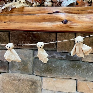 May include: A decorative garland featuring three small, off-white fabric ghost figures suspended on a thin, light-colored twine string. Each ghost has a rounded head with three small, dark, circular markings representing eyes and a mouth.
