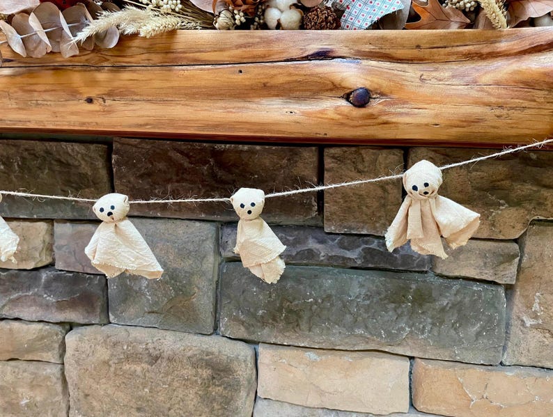 May include: A decorative garland featuring three small, off-white fabric ghost figures suspended on a thin, light-colored twine string. Each ghost has a rounded head with three small, dark, circular markings representing eyes and a mouth.