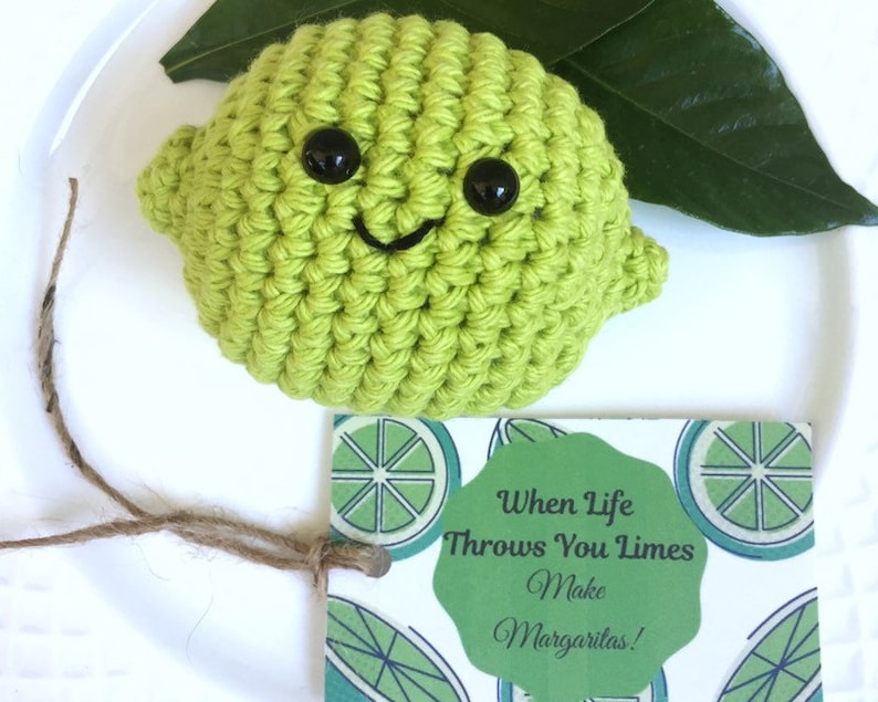 May include: A crocheted lime with a smiling face and googly eyes. The lime is sitting on a white plate with a green leaf and a card that says "When life throws you limes, make margaritas!"