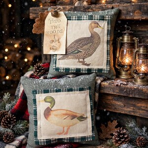 May include: Two square decorative pillows featuring fabric patches with duck illustrations. The top pillow has a beige tag attached with a small bell, printed with the text "INTO THE WOODS I GO!". Both pillows have a green textured base fabric.