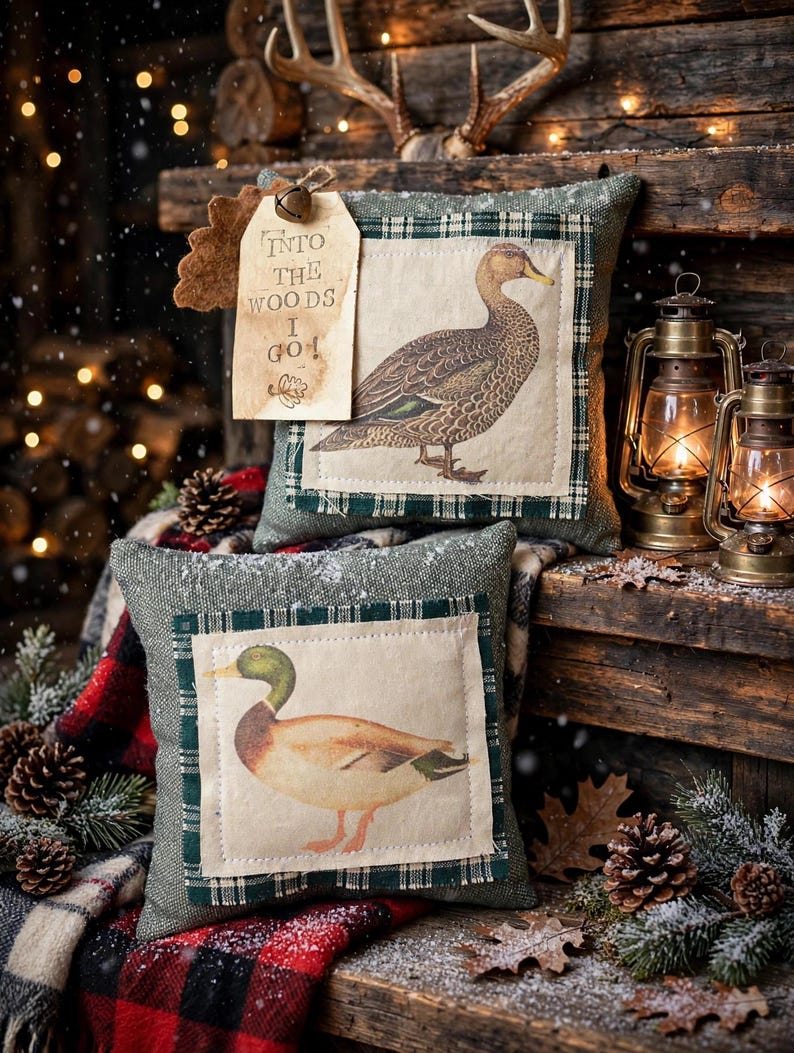 May include: Two square decorative pillows featuring fabric patches with duck illustrations. The top pillow has a beige tag attached with a small bell, printed with the text "INTO THE WOODS I GO!". Both pillows have a green textured base fabric.