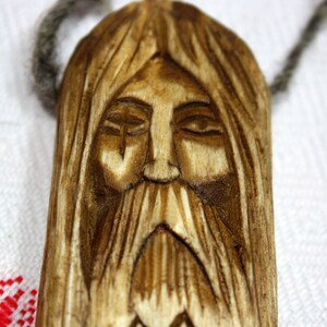 May include: A wooden pendant carved with a detailed face, featuring a long beard and closed eyes. The pendant is brown and has a natural wood grain.