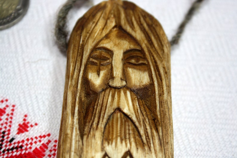 May include: A wooden pendant carved with a detailed face, featuring a long beard and closed eyes. The pendant is brown and has a natural wood grain.