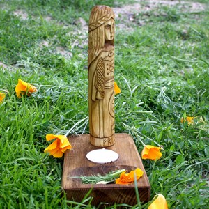 May include: A carved wooden statue of a woman with long hair and a detailed outfit, standing on a wooden base with a candle and sprigs of rosemary. The statue is surrounded by green grass and orange flowers.