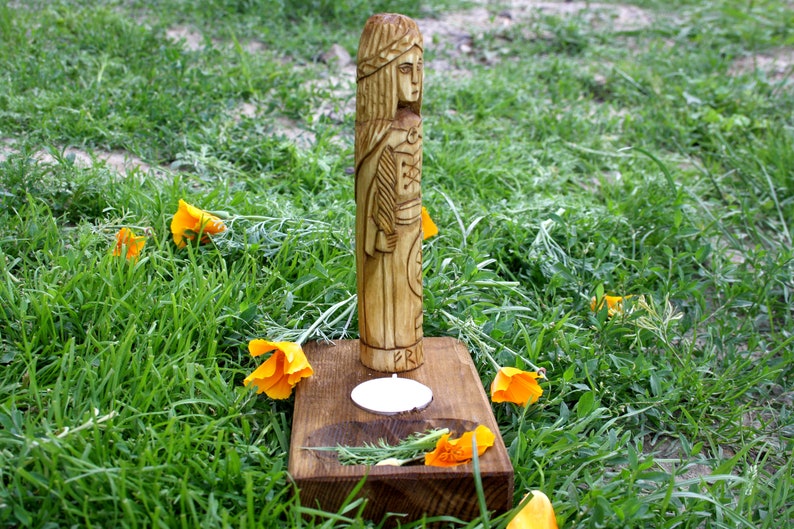 May include: A carved wooden statue of a woman with long hair and a detailed outfit, standing on a wooden base with a candle and sprigs of rosemary. The statue is surrounded by green grass and orange flowers.
