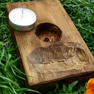 May include: A rustic, wooden candle holder with a natural finish. It features a circular indentation for a tea light candle and a carved, organic shape. The candle holder is set on green grass with orange flowers.